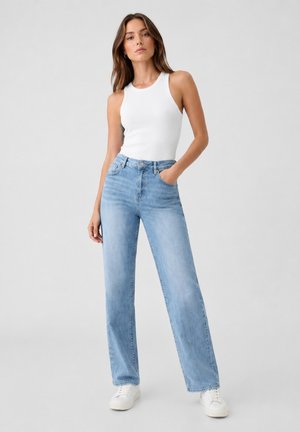 Frau steht, trägt ein weißes ärmelloses Top, hellblaue Jeans mit hoher Taille und weiße Turnschuhe vor einem schlichten grauen Hintergrund.