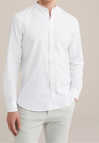 Man med långärmad vit skjorta med mandarinkrage och ljusa byxor, med en hand i fickan.