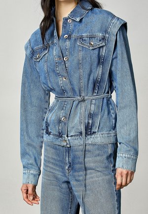 Kvinde iført en lyseblå denimjakke med bælte, foranlommer og lange ærmer, kombineret med matchende denimjeans.