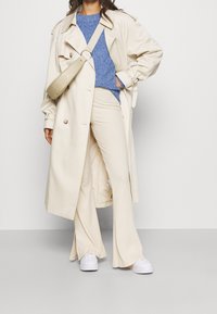 Trench beige, pull en tricot bleu, pantalons larges crème et baskets blanches. Le manteau a un design croisé et une ceinture.