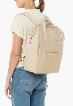 Woman with ponytail wearing cream short-sleeve top and blue jeans carrying beige backpack with zippered front pocket and top handles.