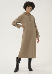 Abito camicia lungo marrone realizzato in tessuto, con colletto, spacchi laterali e due tasche frontali, abbinato a stivali neri fino al ginocchio.