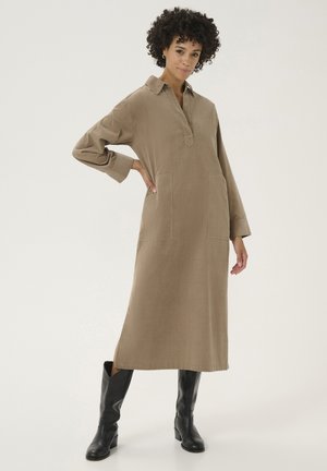 Long brown shirt dress made of fabric with a collar, side slits, and two front pockets, paired with black knee-high boots.