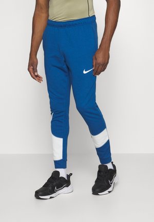 Man draagt blauwe Nike joggers met witte panelen, zwarte Nike sneakers en witte sokken, staand tegen een effen achtergrond.