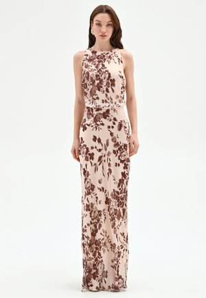 Donna in piedi che indossa un abito beige senza maniche e lungo fino a terra, con motivi floreali marroni, su uno sfondo bianco uniforme.