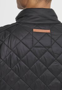 Veste matelassée noire avec col montant, en matériau brillant avec un accent en cuir portant l'inscription "BLEND" au dos.