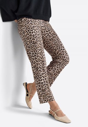 Pantalon slim à imprimé léopard associé à des ballerines slingback pointues beiges et un haut noir, modèle appuyé contre un mur blanc.