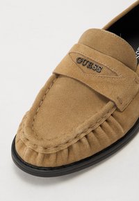Brune semsket loafer med rund tå, plisserte detaljer og en svart gummisåle. Pyntet med en sølv "GUESS"-logo på stroppen.