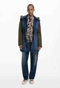 Denim dlhý vest s kapucňou s kožušinkou, zelenými rukávmi a prednými vreckami. V kombinácii s vzorovanou košeľou a voľnými modrými džínsami. Svetlé topánky.
