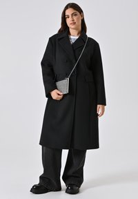 Cappotto di lana nero doppiopetto con ampi risvolti, abbinato a jeans neri a gamba larga e calzature nere chunky; tiene in mano una piccola borsa metallica.