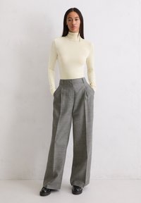 Vrouw die tegen een witte muur staat, gekleed in een crèmekleurige coltrui, hoge grijze wijde broek en zwarte schoenen, met haar handen in haar zakken.