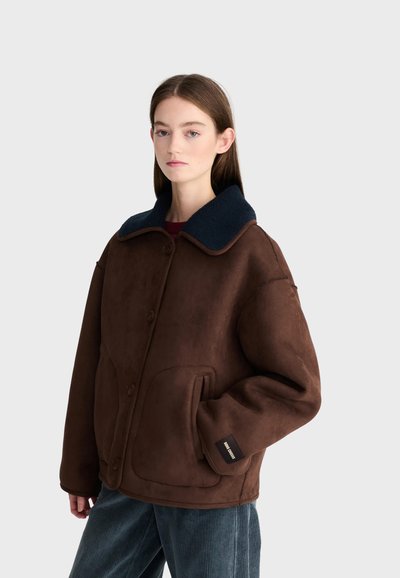 Jeune femme aux longs cheveux bruns portant une veste marron foncé boutonnée avec un col noir et un pantalon en velours côtelé bleu, debout devant un fond gris.