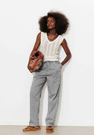 Femme aux cheveux bouclés portant un top en maille blanc sans manches, pantalon gris à cordon, sandales marron, tenant un sac marron plié à motif.