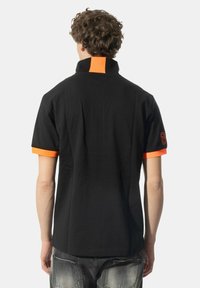 Maglietta polo nera con colletto alto, dettagli arancioni sul colletto e sulle maniche, e il numero 6 ricamato sulla manica sinistra.