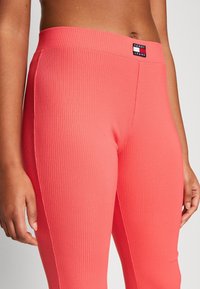 Zbliżenie na osobę noszącą jasne koralowe legginsy ze ściągaczem i naszywką z logo Tommy Jeans na pasku, z rękami swobodnie opadającymi wzdłuż boków.