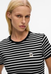 Blonde woman with freckles wearing a black and white striped t-shirt featuring a small cartoon face with sunglasses embroidered on the chest.