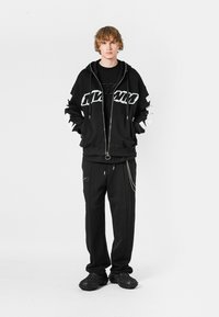 Sudadera negra con cremallera y acentos gráficos en blanco, superpuesta sobre una camiseta negra. Combinada con pantalones negros con una cadena y zapatos negros texturizados.