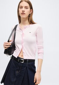 Νέα γυναίκα που φοράει ροζ ανοιχτό ριγέ ζακέτα με κουμπιά, σκουρό μπλε πλισέ φούστα με μαύρη ζώνη, κρατώντας μια μικρή μαύρη τσάντα στον ώμο.