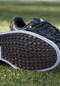 Črne športne copate z teksturirano gumijasto podplatom z valovitimi vzorci, belim srednjim podplatom in črnimi vezalkami, postavljene na zeleno umetno travo.