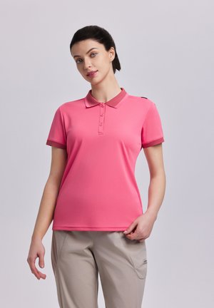 Vrouw met donker haar naar achteren gebonden, gekleed in een roze poloshirt met korte mouwen en beige broek, staande tegen een lichtgrijze achtergrond.