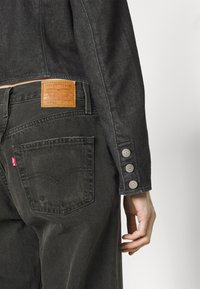 Veste en denim noir à coupe courte, avec trois boutons en métal argenté sur la manche, et jean noir avec une étiquette en cuir marron à l'arrière.