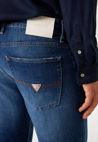 Temne modri denim kavbojke z Guess logotipom na hrbtu, ki ima trikotno zasnovo, subtilno šivanje in teksturirano tkanino.