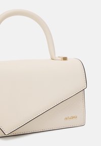 Kremowa torebka o usztywnionym kształcie, z fakturowanym panelem, kontrastującymi ciemnymi obszyciami oraz złotym logo.
