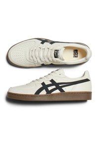 Hvide lædersneakers med sorte accenter, der har en perforeret tå, flade snørebånd og en brun gummisål. Asics-logoet på tungen.