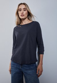 T-paita, jossa on pitkät hihat, laaja pääntie ja raglan-hihat, tumman sininen, yhdistettynä farkkuihin, joissa on koristeellisia yksityiskohtia lahkeissa.