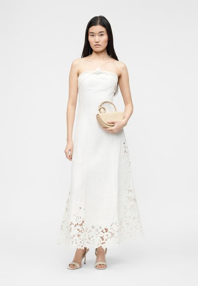 Γυναίκα με μακριά σκούρα μαλλιά, φορώντας λευκό φόρεμα χωρίς τιράντες με δαντέλα στο τελείωμα, μπεζ ψηλοτάκουνα και κρατώντας μικρή μπεζ τσάντα με στρογγυλή λαβή.