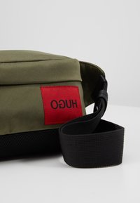 Torba na ramię z oliwkowozielonego materiału z czarnym regulowanym paskiem oraz czerwoną prostokątną naszywką z napisem "HUGO" czarnymi literami.