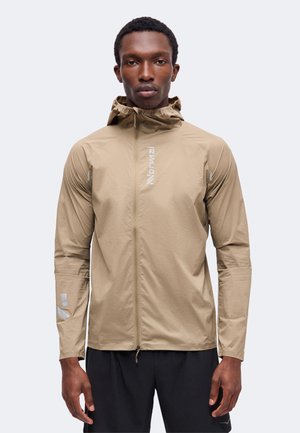 Homme portant une veste légère beige à capuche avec fermeture éclair à l'avant et logo réfléchissant, debout les bras détendus devant un fond uni.