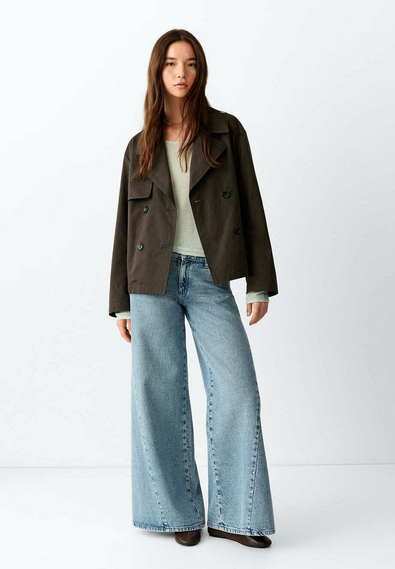 Jeune femme portant une veste croisée marron foncé, un pull vert clair, un jean bleu clair à jambes larges et des chaussures foncées, debout sur un fond blanc.
