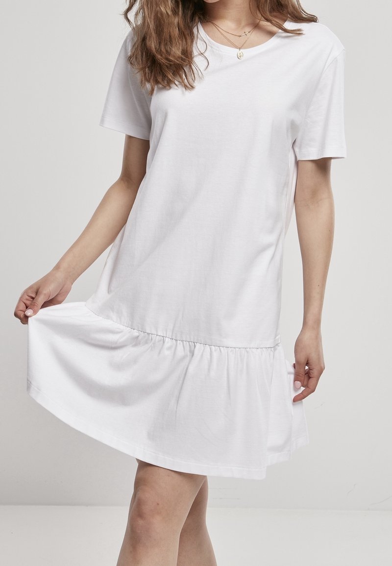 Žena v krátkých rukávech bílých šatech se staženým lemem, držící sukni mírně od těla, na jednobarevném světlém pozadí.