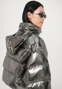 Metāliska pūkaina jaka ar kapuci, ar spīdīgu, teksturētu virsmu un šķērsgriezuma daļām. Tumšas, minimālistiskas saulesbrilles papildina izskatu.