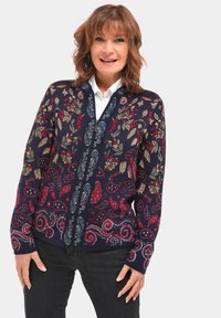 Marineblauwe cardigan met bloemen- en paisleypatronen in rood, groen en blauw. Voorzien van een ritsluiting en lange mouwen, gemaakt van een gestructureerde breimateriaal.