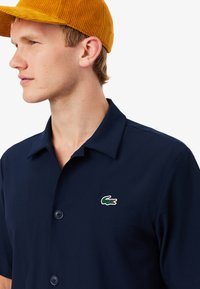 Polo shirt blu navy a maniche corte con una texture morbida e due bottoni frontali. Presenta un piccolo logo Lacoste verde e rosso sul petto.