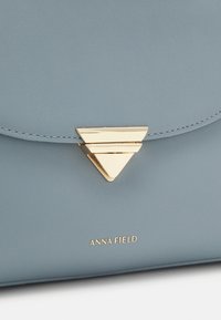 Bolso de mano de imitación cuero azul claro con un cierre triangular dorado y el logo en relieve "ANNA FIELD" en la parte inferior. Textura suave y diseño elegante.