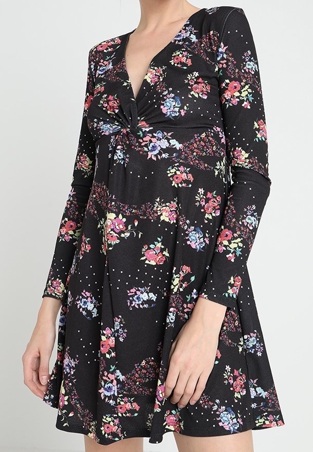Femme portant une robe noire à manches longues avec un nœud torsadé à l'avant et un motif floral multicolore, debout devant un fond uni.