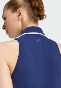 Námornícka polo košeľa s textúrovanou tkaninou, vysokým golierom a bielymi akcentmi. Má malé modré logo Adidas na chrbte.