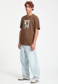 Brun bomulds T-shirt med et grafisk logo design på forsiden, parret med lyseblå afslappede jeans og sort-hvide sneakers.
