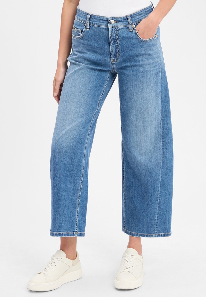 Personne portant un jean large court bleu clair avec poches avant et arrière et des baskets blanches sur un fond uni.