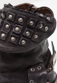 Bota de piel negra con superficie texturizada, con tachuelas cuadradas y remaches plateados redondos. Acentuada con una correa y detalle de hebilla.