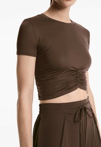 Top corto marrone con maniche corte, caratterizzato da dettagli arricciati nella parte anteriore. Realizzato in tessuto elasticizzato, abbinato a pantaloni a vita alta dello stesso colore.