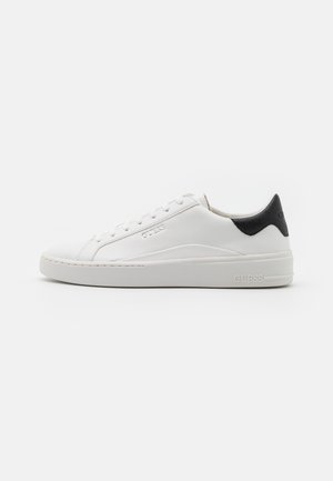Witte lage sneaker met witte veters, dikke zool, zwart detail op de hiel en een reliëf GUESS-logo aan de zijkant. Tegen een effen achtergrond.
