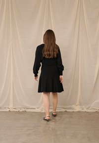 Moteris su ilgais rudas plaukais, vilkinti juodą suknelę ir zebra rašto aukštakulnius, stovinti ant betono grindų priešais smėlio spalvos užuolaidas.