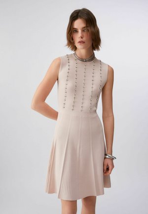 Jauna sieviete valkā bezroku bēša krāsas adītu kleitu ar vertikālu pērlīšu rotājumu un plisētu svārku daļu, papildinot izskatu ar sudraba aprocēm un kaklarotu.