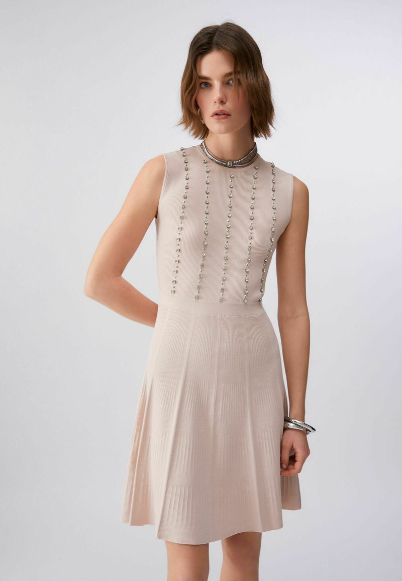 Jeune femme portant une robe beige en maille sans manches avec des détails verticaux en perles et une jupe plissée, accessoirisée avec des bracelets argentés et un collier ras du cou.