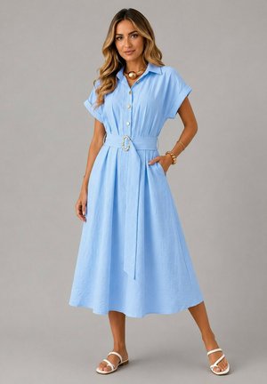 Femme portant une robe midi bleu clair ceinturée à manches courtes et des sandales blanches, debout avec une main dans la poche sur un fond gris.