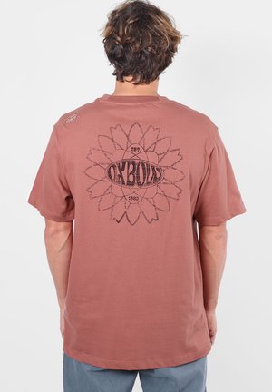 Personne aux cheveux courts et ébouriffés portant un t-shirt marron avec un grand motif floral et le texte "OXBOW" dans le dos.